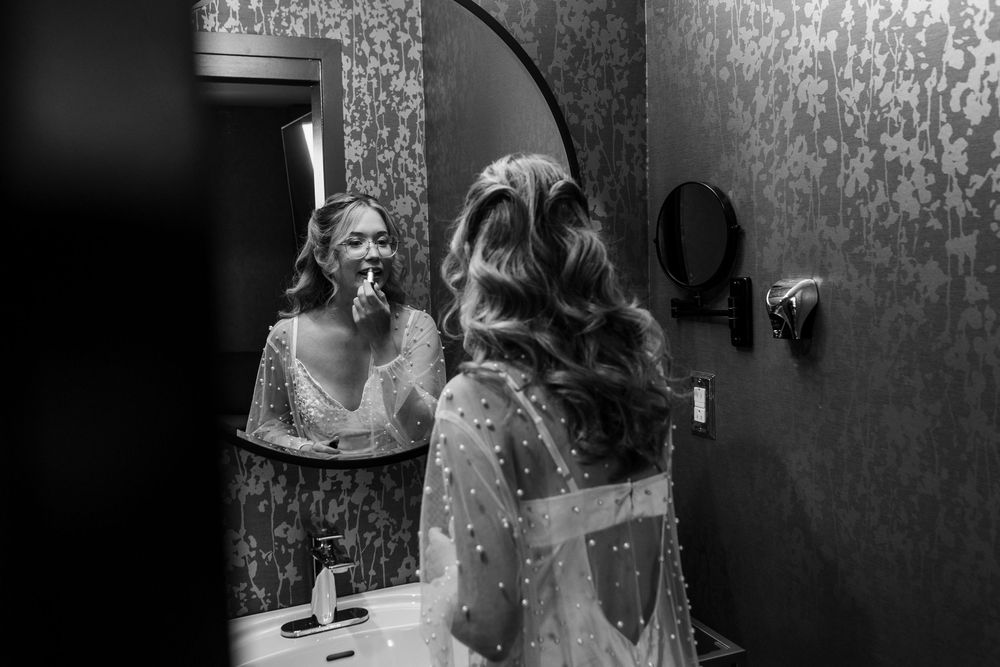 Black and white photo of a bride touching up her makeup in a decorative bathroom mirror before her wedding.