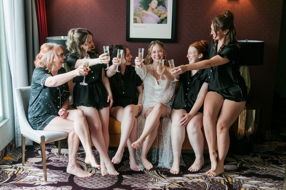 Bride and bridesmaids in black pajamas toasting with champagne glasses during wedding morning preparations.