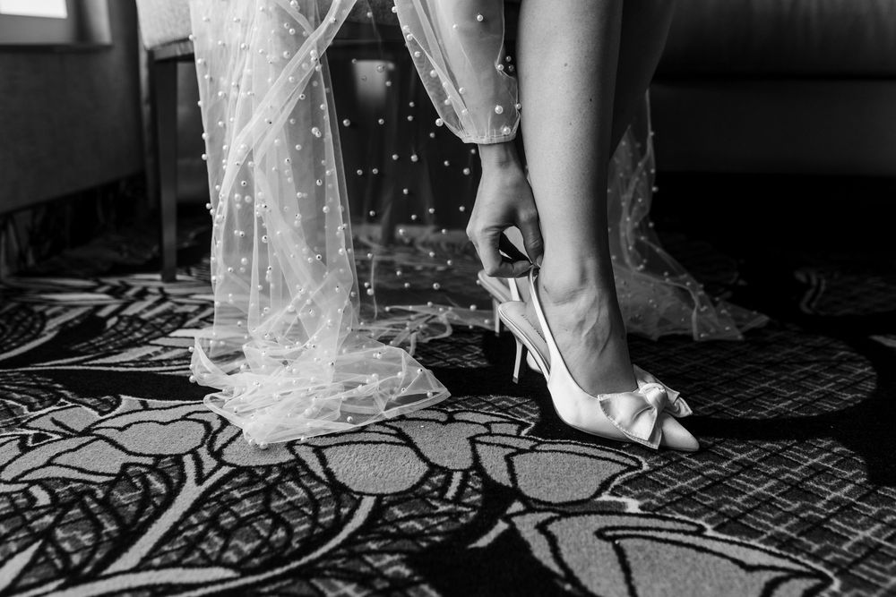 Bride putting on white satin bow heels under polka dot tulle wedding dress in black and white photo.