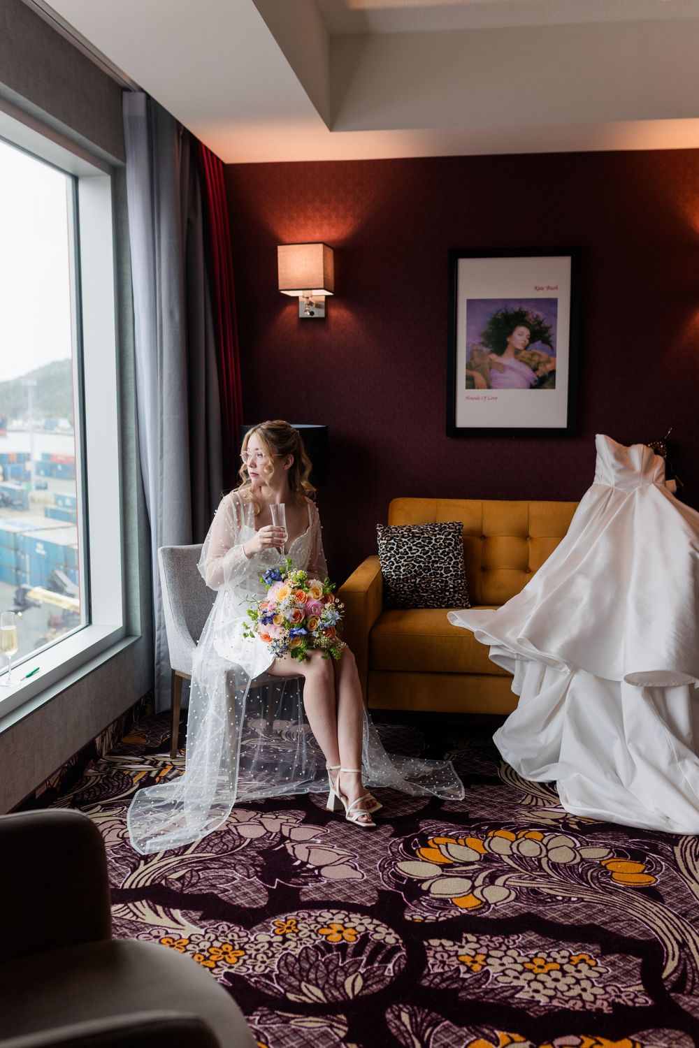 Bride in wedding dress sits by window holding colorful bouquet in elegant hotel room with yellow sofa.