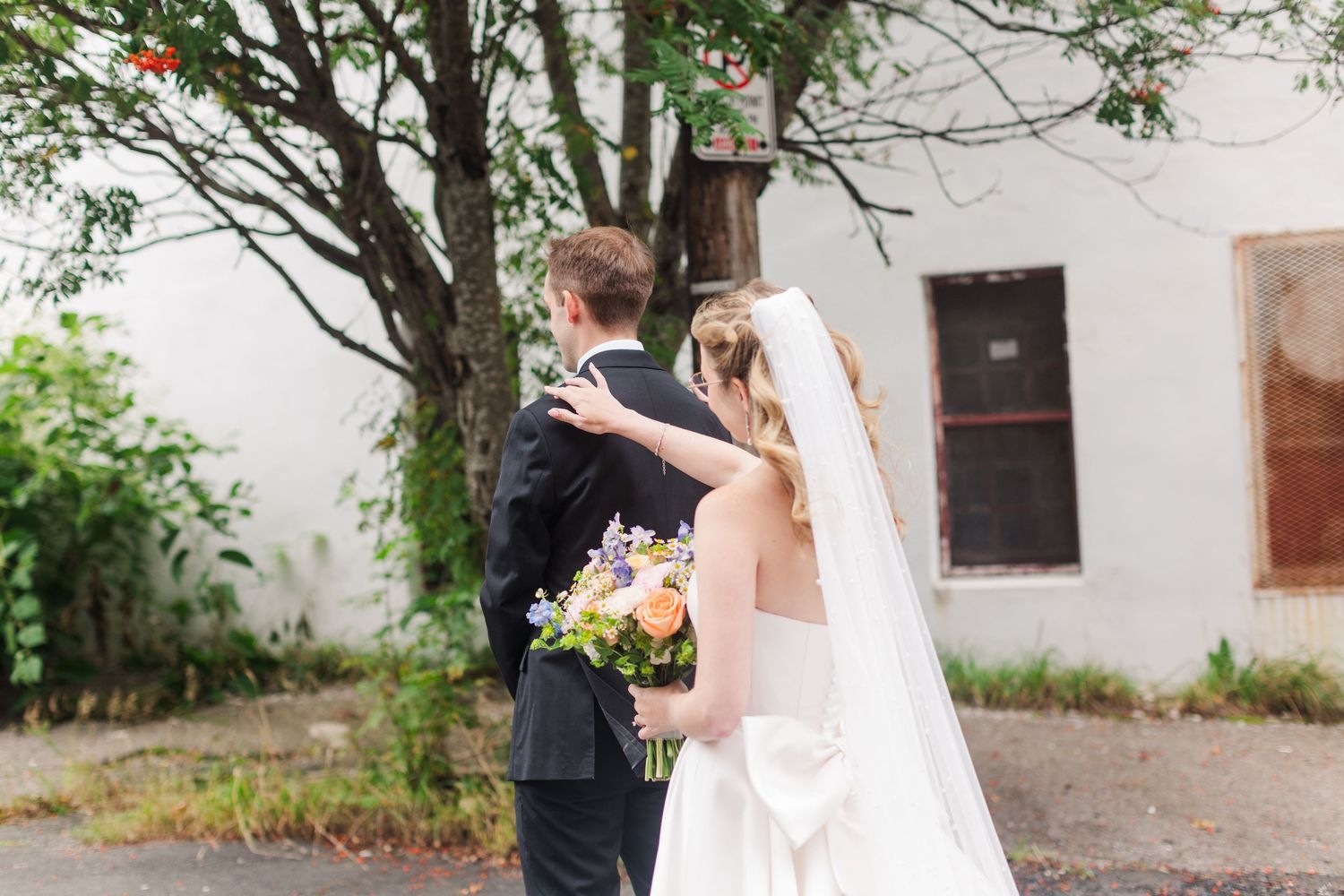 Bride in white gown and veil touches groom's shoulder outdoors near tree, holding colorful bouquet.