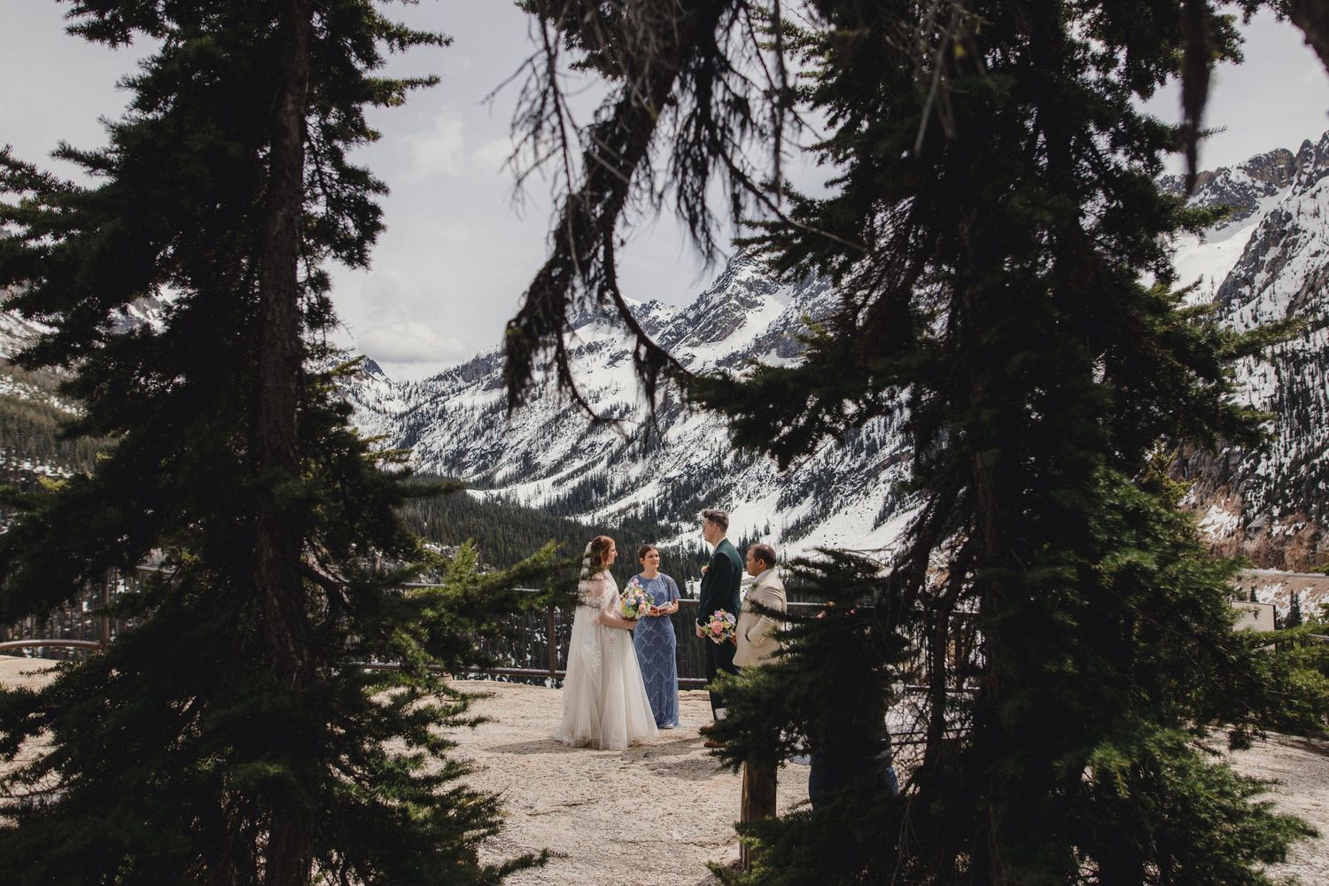 Couple exchanges vows outdoors surrounded by tall pine trees with snow-capped mountains in the background in north cascades in washington.