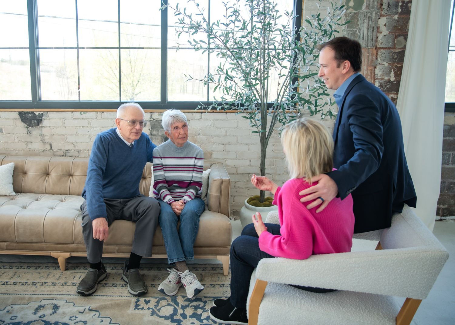 Two elderly people sit on a sofa while a man in a suit and a woman in pink stand nearby in a modern living room.