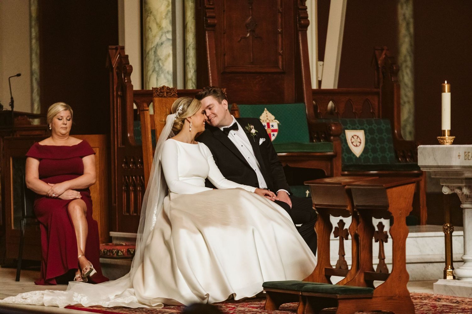 Bride and groom share an intimate moment seated at church altar during wedding ceremony, with guest in red dress seated behind.