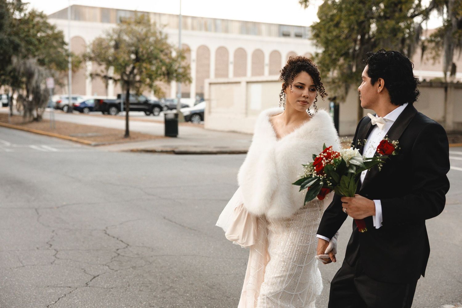 Bride in white gown with fur wrap holds bouquet beside groom in tuxedo crossing a street on their wedding day.
