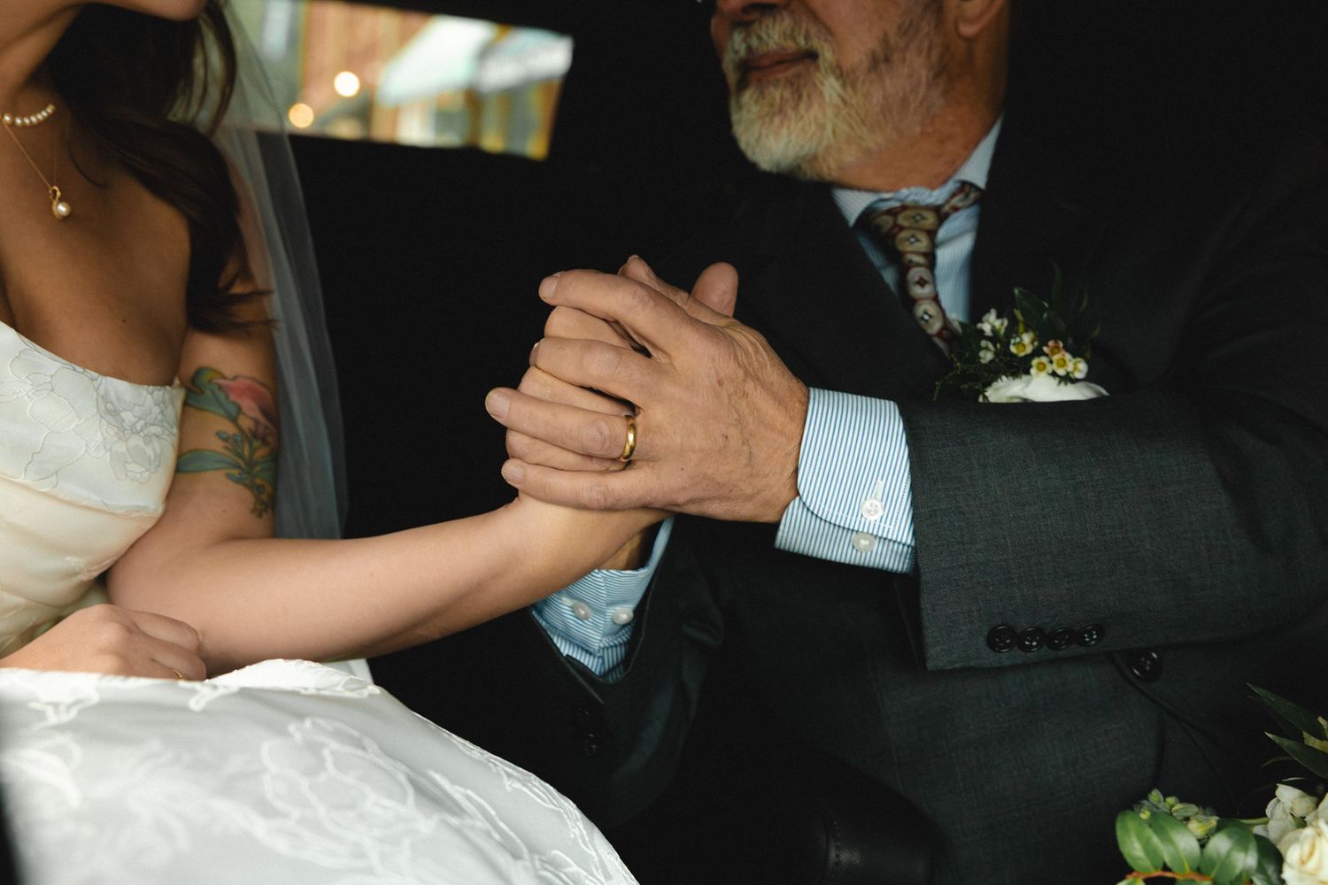 Bride and older gentleman holding hands tenderly in a car, wearing wedding attire with floral boutonniere.