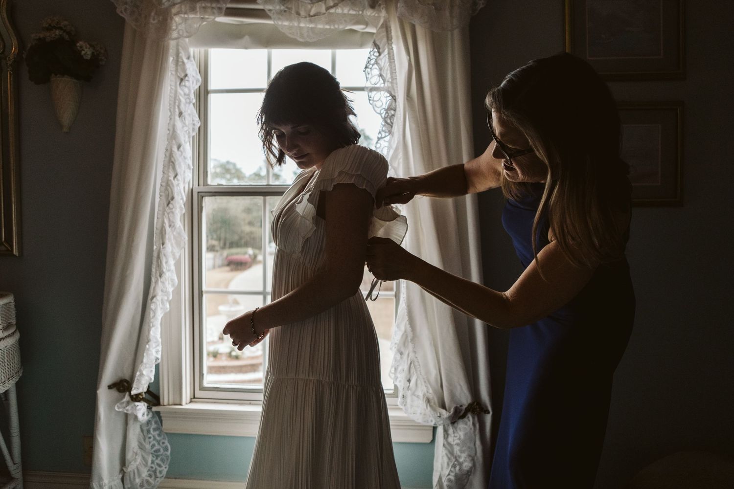 Bridesmaid in blue dress helping bride with her white wedding gown near a sunlit window indoors.