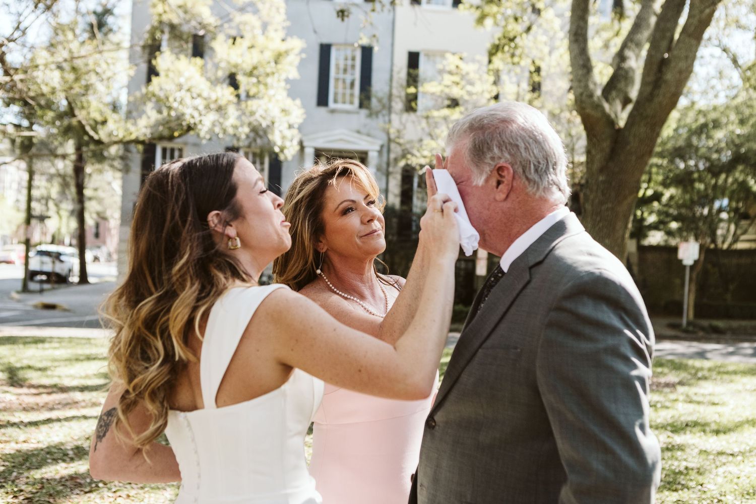 A bride adjusts makeup on an older man while a woman in pink looks on outdoors near a historic building.