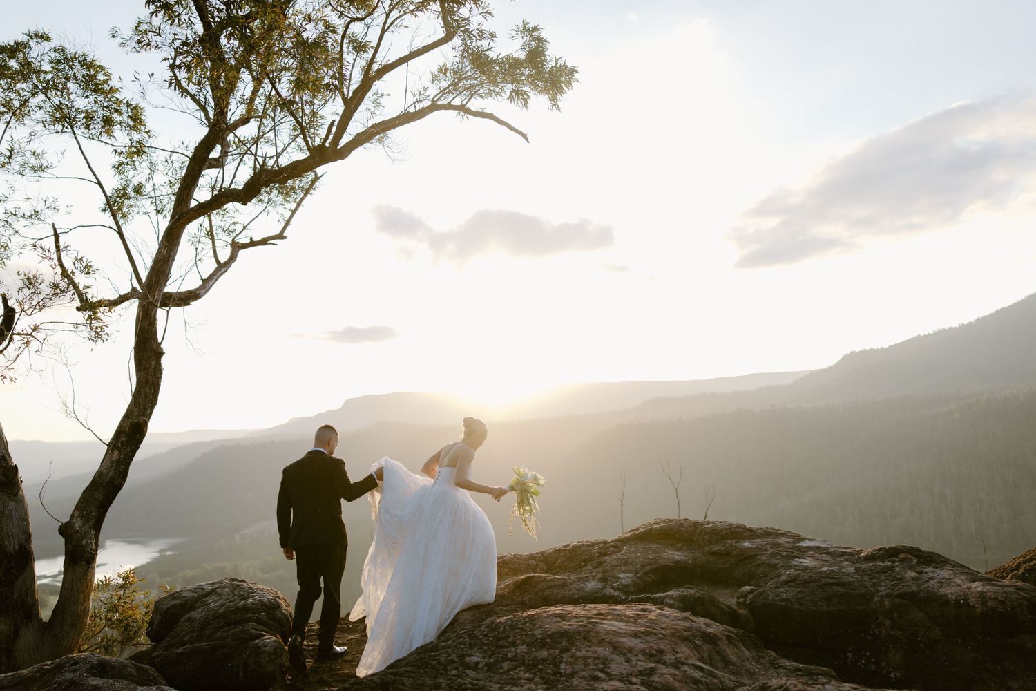 A laughing bride in a short white dress holds hands with a groom in a black tuxedo on a green farm lawn.