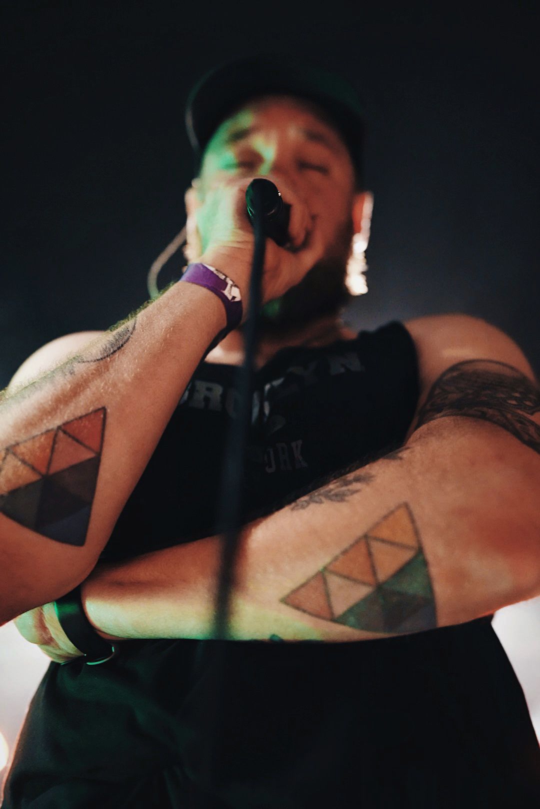 Tattooed performer singing into microphone on stage with triforce tattoos visible on both arms under green and purple lights.