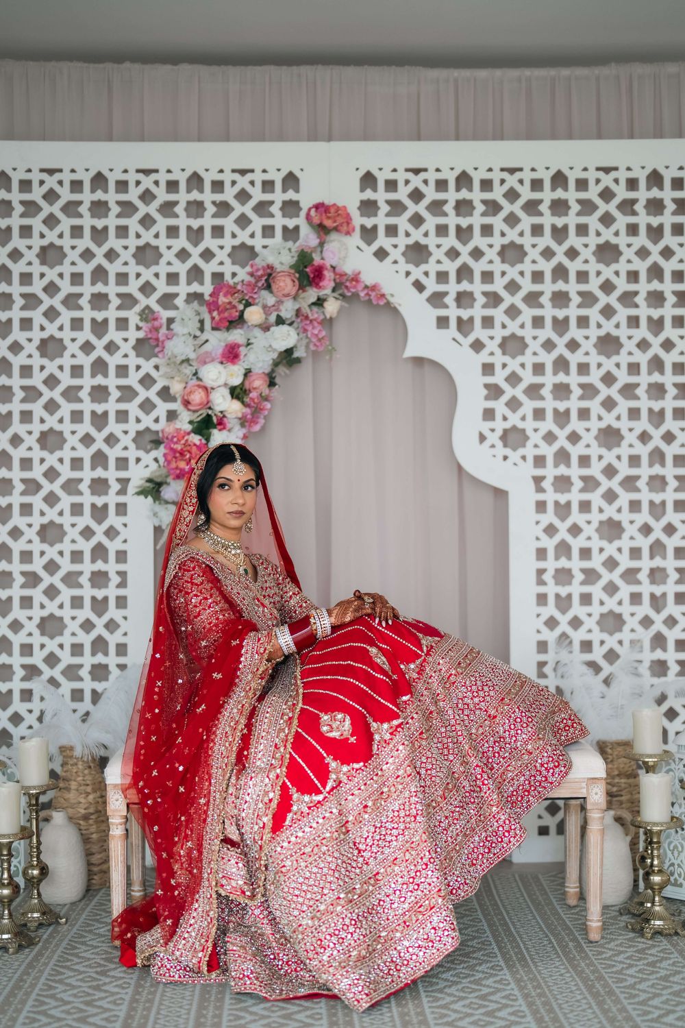 Bride in red embroidered lehenga with dupatta poses before white lattice backdrop with pink floral arch.
