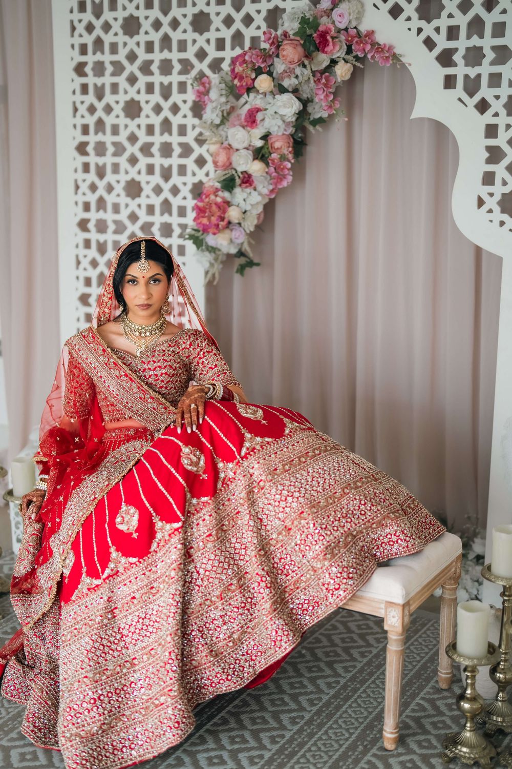 Indian bride in red embroidered lehenga choli with gold jewelry sits elegantly before floral backdrop.