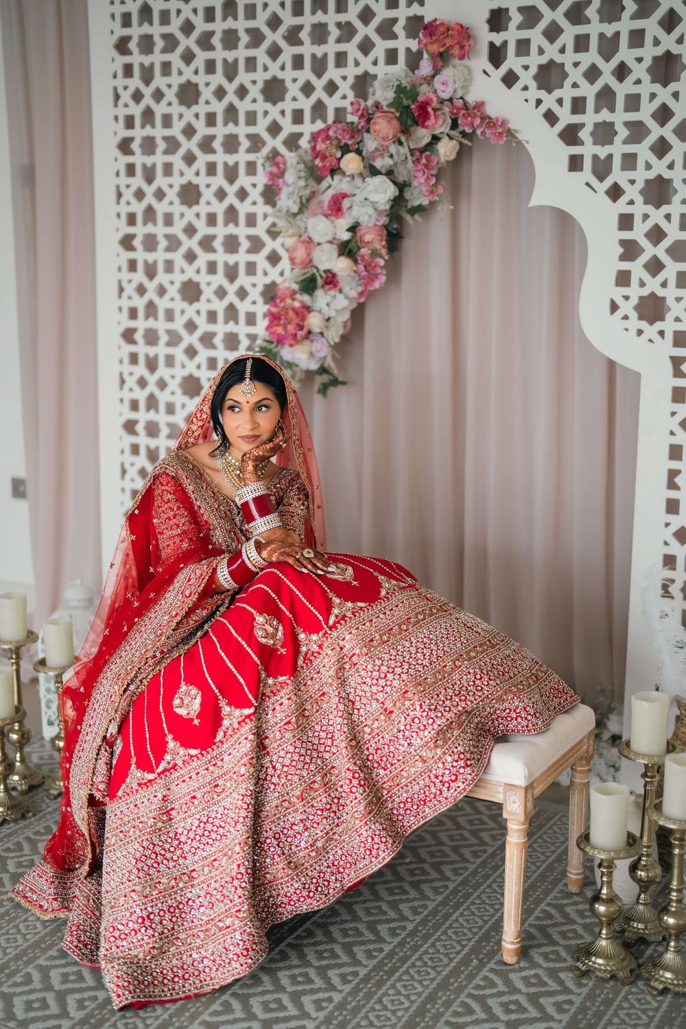 Indian bride in red lehenga with gold embroidery sits elegantly before floral backdrop and white lattice wall.