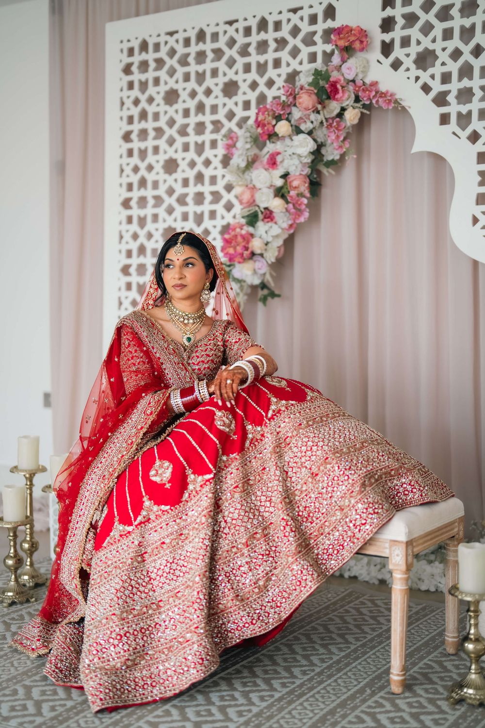 Indian bride in red embroidered lehenga choli with gold jewelry sits elegantly before floral backdrop.