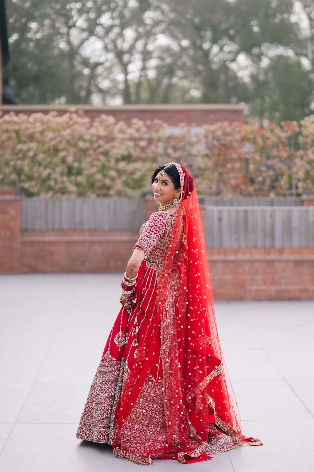 Indian bride in red embroidered lehenga with dupatta smiling outdoors at wedding venue.