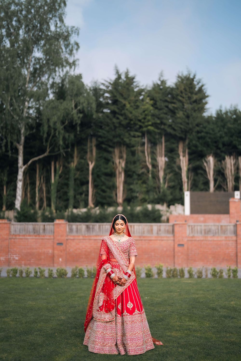 Bride in red embroidered lehenga choli with dupatta stands on green lawn with trees and brick wall backdrop.