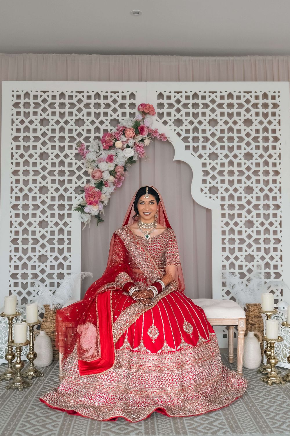 Indian bride in red lehenga with gold embroidery sits before ornate white lattice backdrop with floral arch and candles.