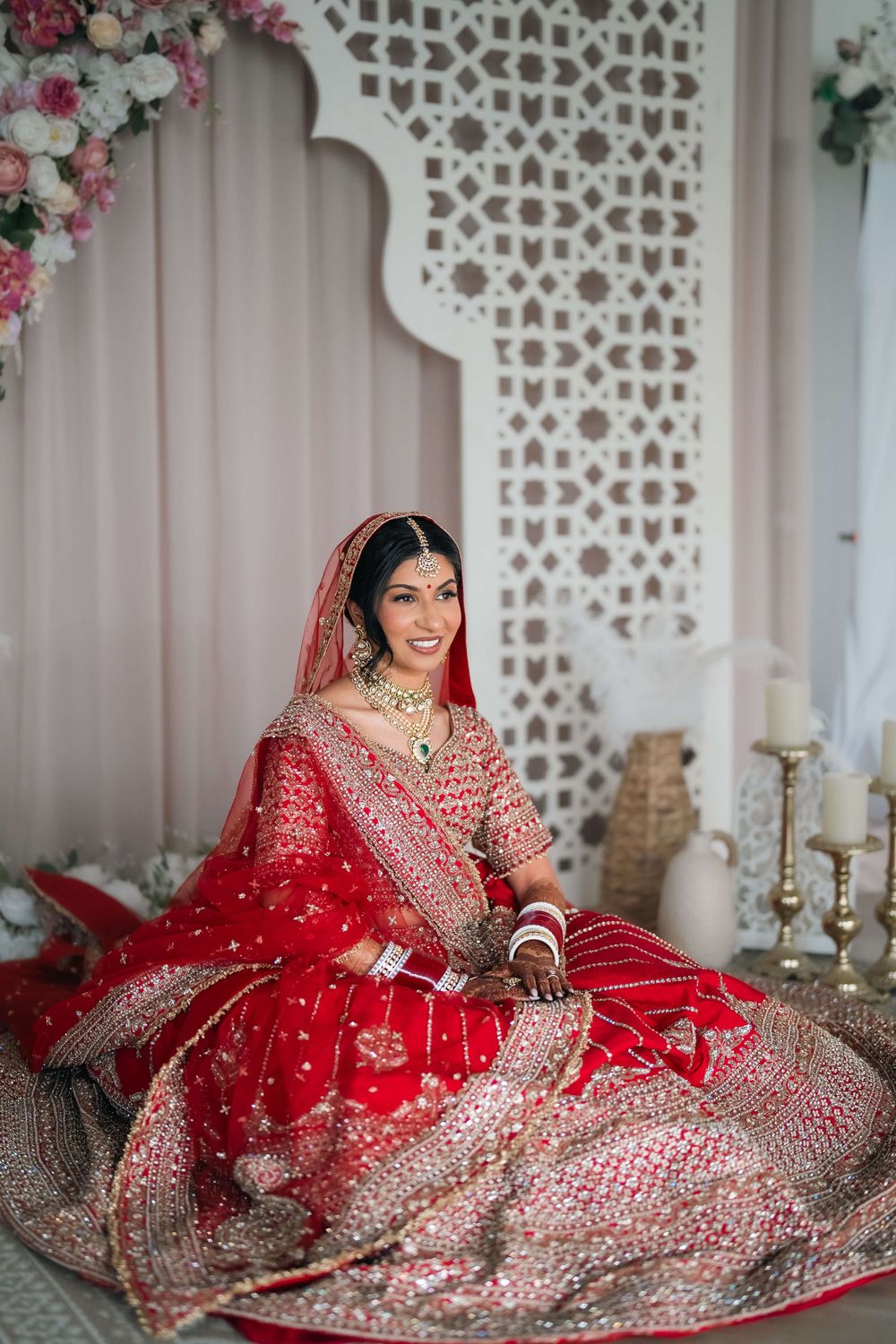 A smiling Indian bride in a red embroidered lehenga and dupatta sits gracefully in a decorated wedding setting.