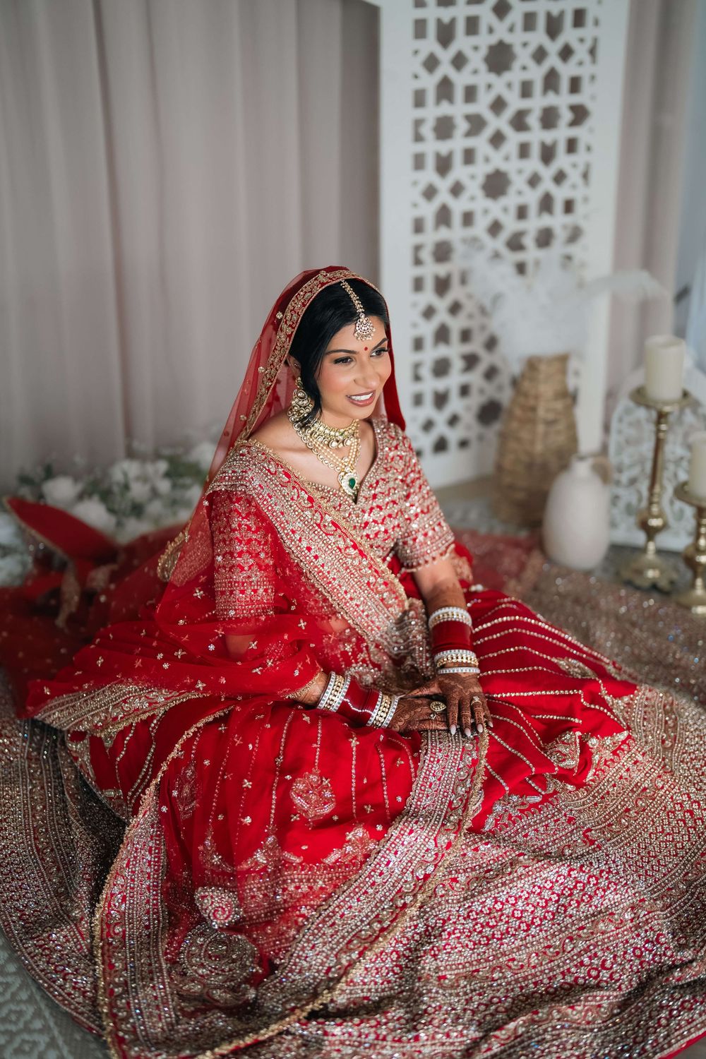 A bride in a red embroidered lehenga with gold jewelry and dupatta sits gracefully in a traditional Indian bridal look.