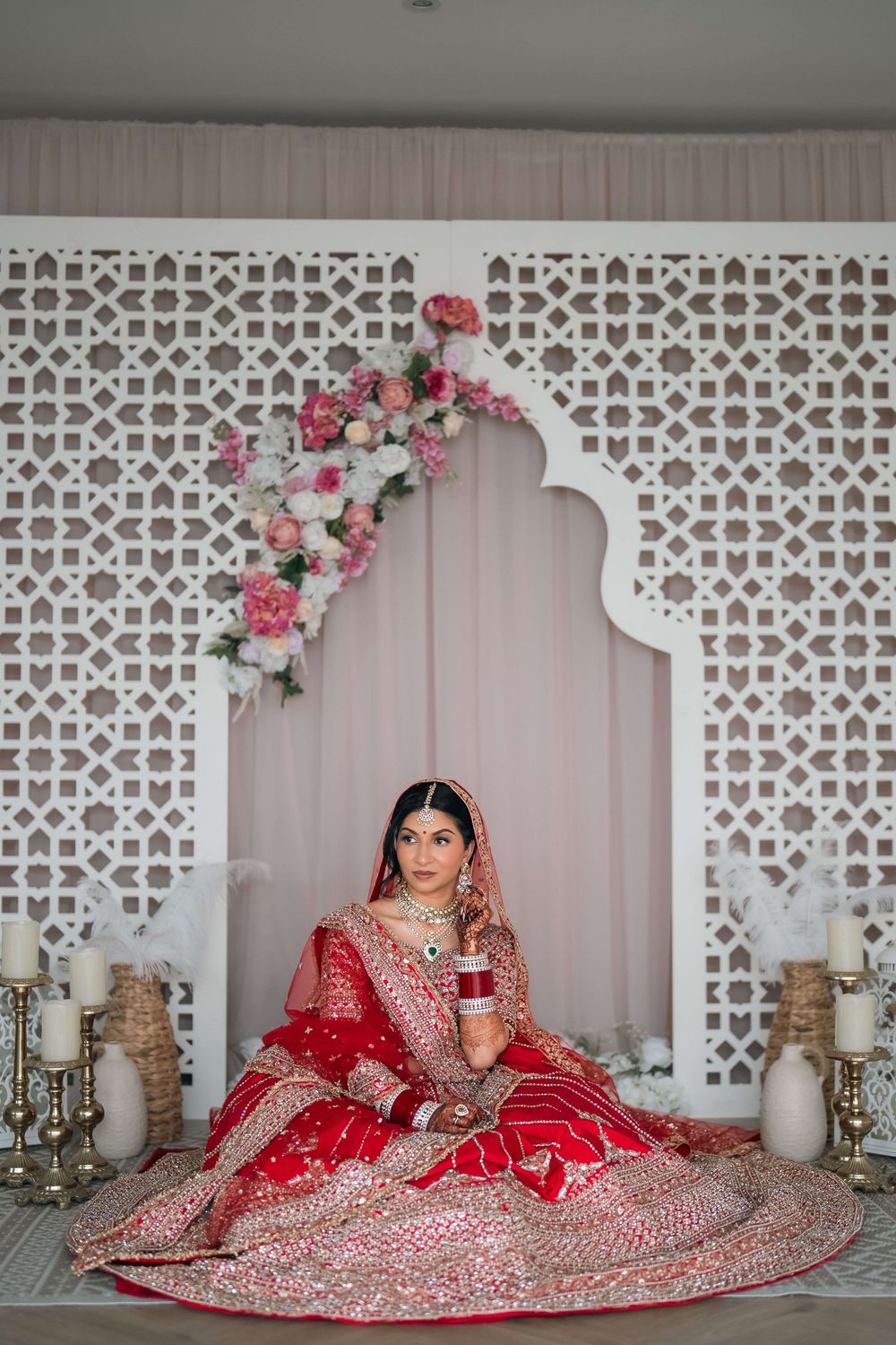 Indian bride in red lehenga sits before ornate white lattice backdrop with floral arch and candles.