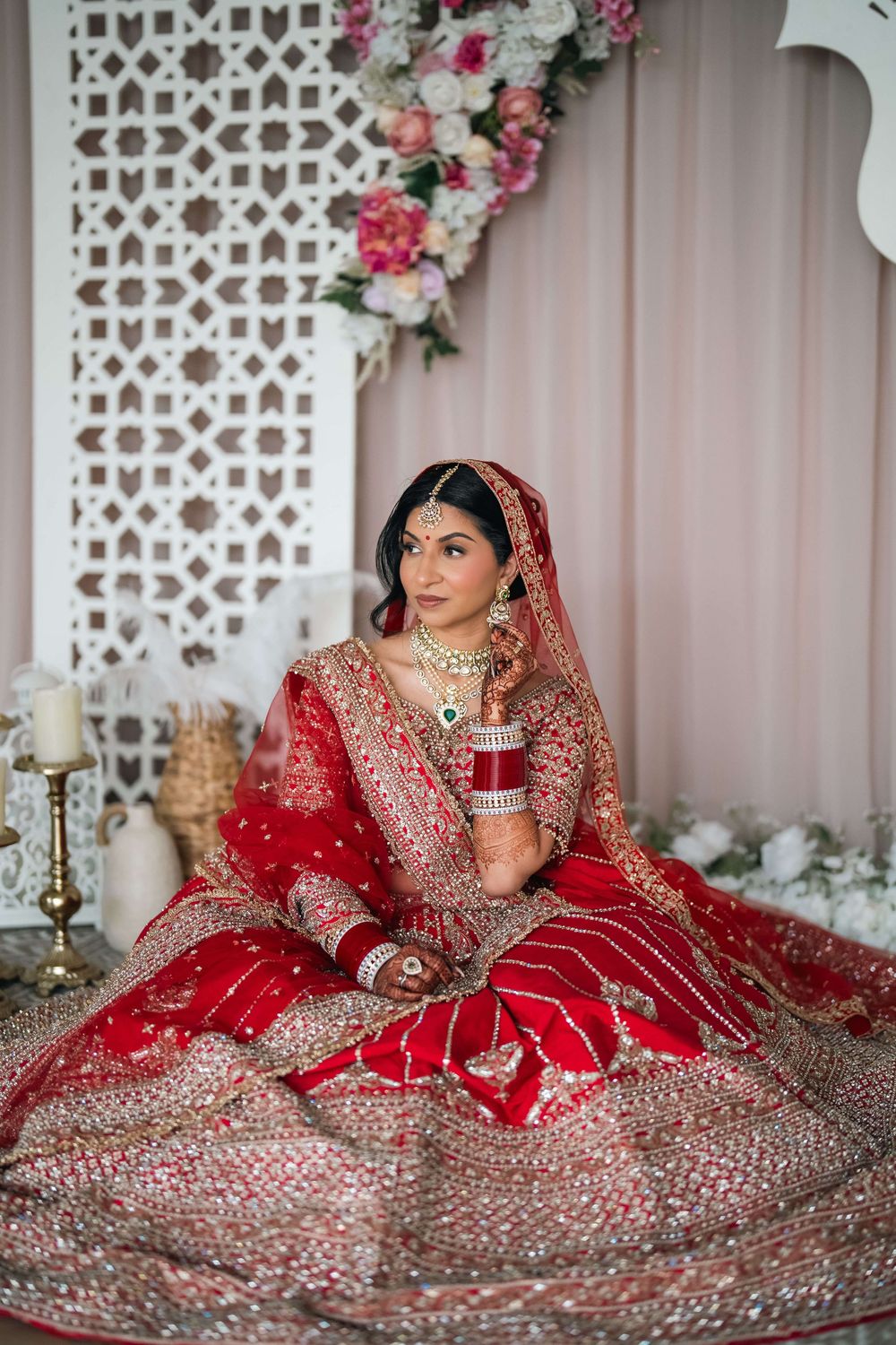Indian bride in red bridal lehenga with gold embroidery, wearing traditional jewelry, seated against floral backdrop.