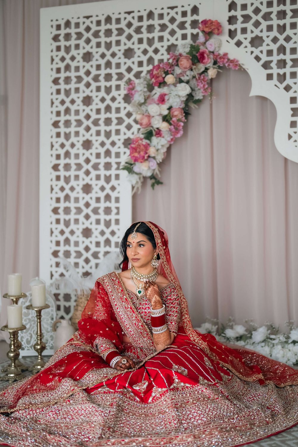 Indian bride in red lehenga with gold embroidery sits gracefully before white lattice backdrop adorned with pink flowers.