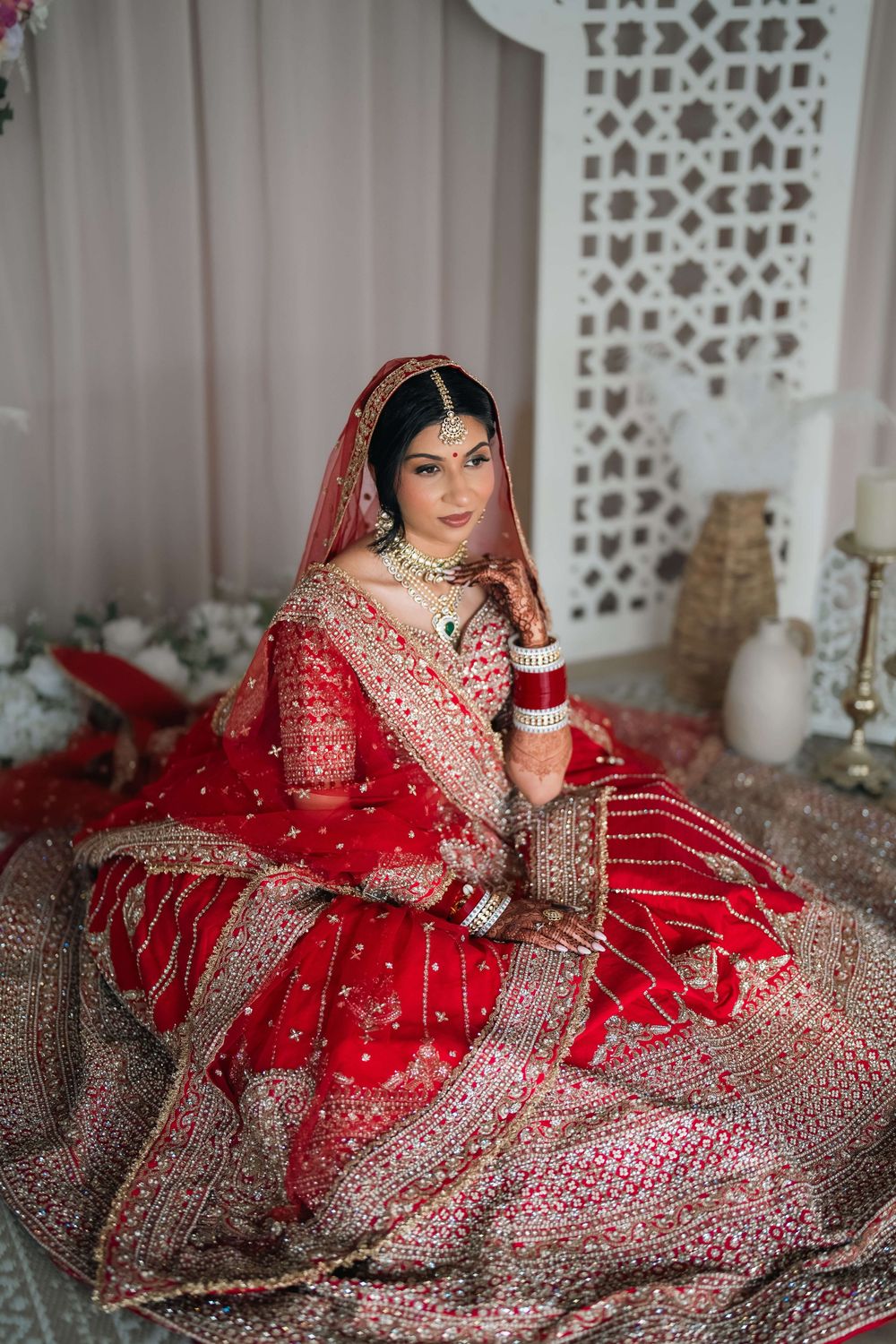 Indian bride in red lehenga with gold embroidery, bridal jewelry, and dupatta, posing elegantly indoors.