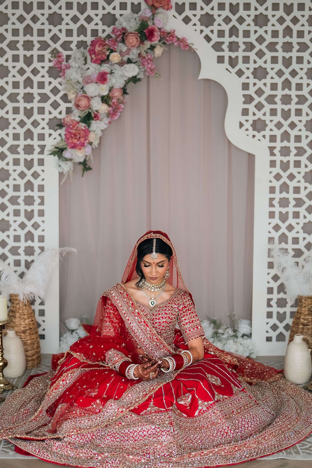 A bride in a red embroidered lehenga sits gracefully before a floral arch backdrop during an Indian wedding ceremony.