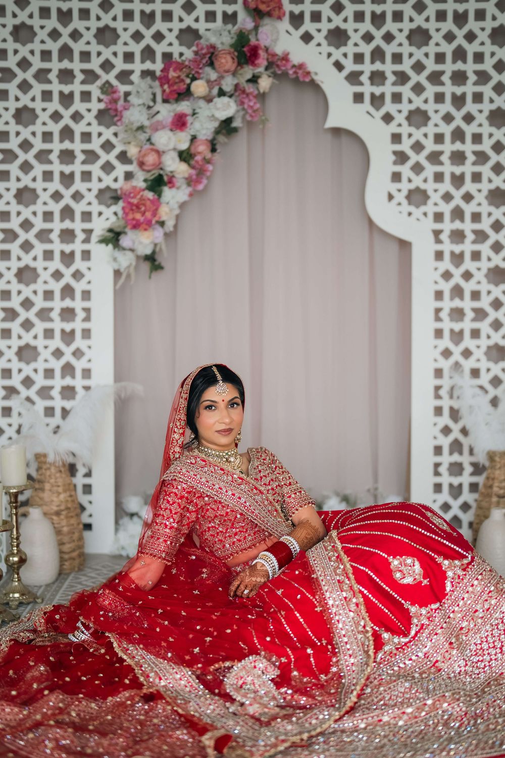 Indian bride in red embroidered lehenga with gold jewelry sits elegantly before ornate white lattice backdrop with pink florals.