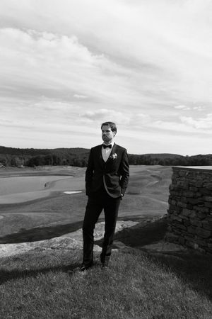 Groom in tuxedo with bow tie stands on stone terrace overlooking golf course in black and white wedding photo.