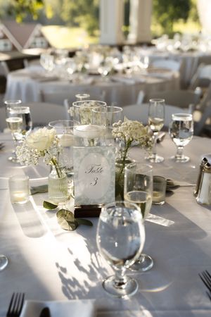 Elegant wedding reception table setting with white floral centerpieces, candles, and a Table 3 sign on white linen.