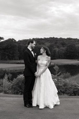 Black and white wedding portrait of bride and groom holding hands, smiling at each other outdoors near a pond.