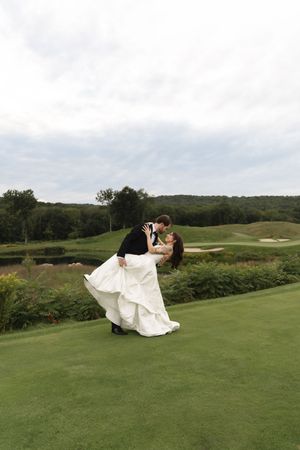 Bride and groom share a romantic dip kiss on a lush green golf course with rolling hills in the background.