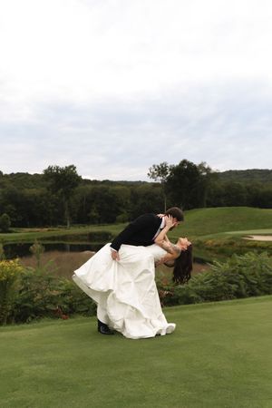 Groom dips bride in white gown for romantic kiss on lush green golf course with scenic pond backdrop.