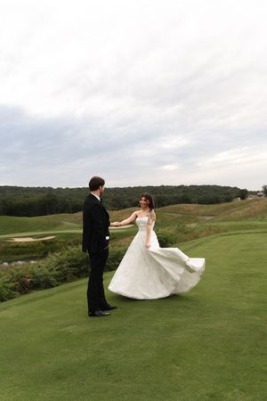 Bride in white gown twirls holding grooms hand on lush green golf course under cloudy sky.