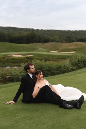 Bride and groom sitting on lush green golf course lawn in wedding attire, with scenic hills and sand traps in background.