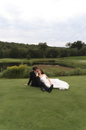 Bride and groom sharing a romantic kiss while sitting on lush green grass at an outdoor wedding venue.