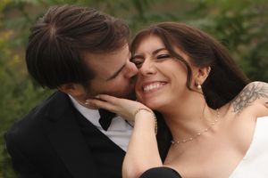 A groom in a black tuxedo kisses his smiling bride with red hair and a tattoo on her shoulder outdoors.
