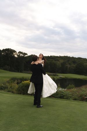 Groom lifts joyful bride in white wedding gown on golf course green with pond and trees in background.