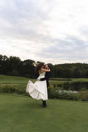 Groom lifting bride in white wedding dress on green lawn with trees and pond at outdoor wedding venue.