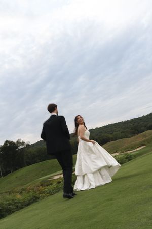 Bride in layered white dress smiles at groom in black tuxedo on scenic golf course fairway.