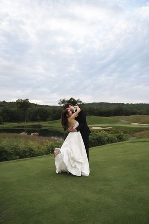 Bride and groom share romantic kiss embrace on green golf course with pond and trees behind.