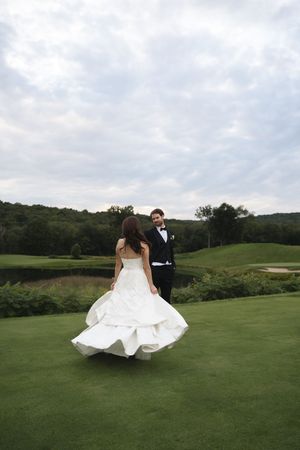 Bride in flowing white gown twirls toward smiling groom on green golf course at outdoor wedding.