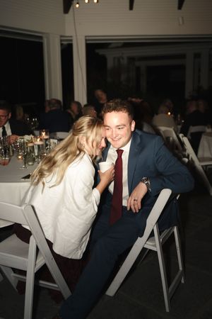 A smiling man in a navy suit with burgundy tie sits at a wedding reception table while a blonde woman whispers in his ear.