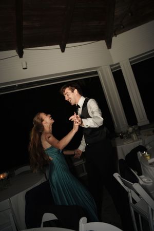 A couple shares a joyful dance at a wedding reception, the woman in a teal gown and man in a black vest.