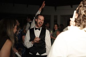 Joyful man in black tuxedo vest raising his hand and laughing while celebrating at a wedding reception.