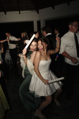 Two women dancing joyfully with foam glow sticks at a lively wedding reception dance floor.