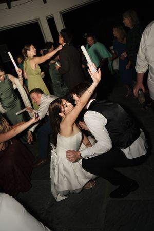 Bride and groom dancing joyfully at wedding reception with guests holding glow sticks on dance floor.