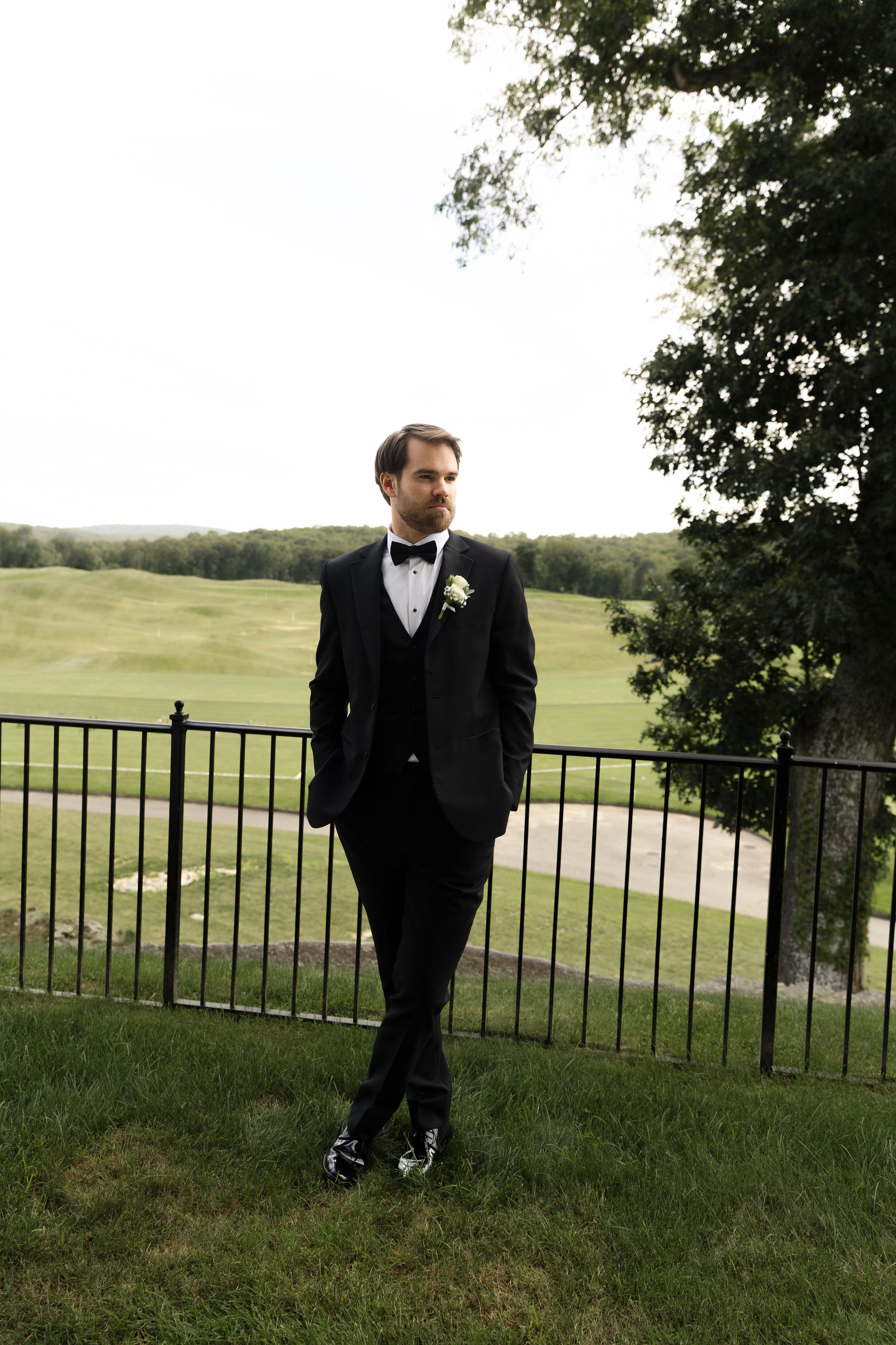 Groom in black tuxedo with bow tie and boutonniere stands by iron fence overlooking green golf course.