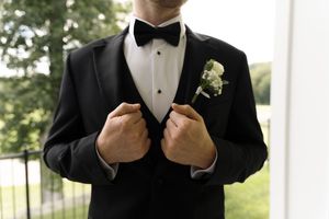 Groom in classic black tuxedo with white boutonniere adjusting his jacket lapels before the wedding.